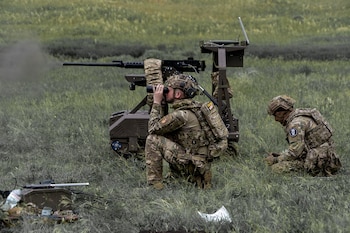 Dos soldados con uniforme de camuflaje en un campo de hierba; uno mira a través de binoculares junto a un sistema de arma robotizada que emite humo por su cañón