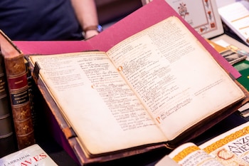 Un libro antiguo abierto con páginas amarillentas y escritura a mano en tinta negra y roja. Se observan otros libros encuadernados al fondo