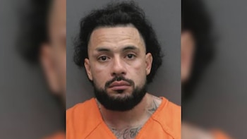 Fotografía policial frontal de un hombre de cabello oscuro y barba con uniforme naranja frente a un fondo gris