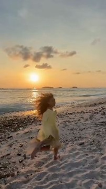 Una persona con cabello rizado y vestimenta clara corre sobre la arena de una playa durante el atardecer. El sol se refleja en el agua del mar