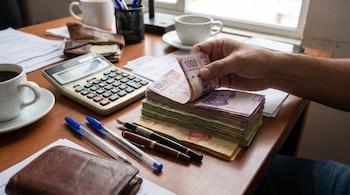 Una mano de hombre contando billetes mexicanos de 1000 pesos sobre una mesa de madera con una calculadora, plumas, documentos y tazas de café.