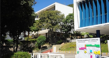 Vista exterior del Colegio Humboldt Caracas, con un edificio blanco y lamas azules en primer plano, rodeado de árboles y vegetación bajo un cielo azul claro