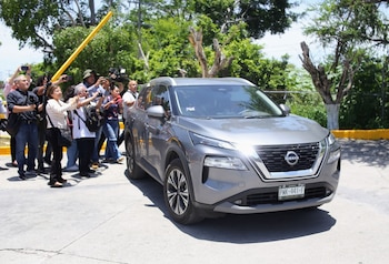 SUV gris con placa de Morelos. Grupo de periodistas con cámaras y teléfonos en una salida al aire libre, con árboles y una barrera amarilla