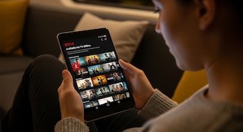Una persona sostiene una tablet que muestra la aplicación de Netflix, con la sección "Disponible para TV sin conexión" y una cuadrícula de miniaturas de títulos.