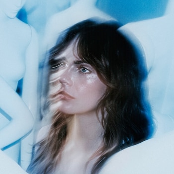 Primer plano de Cara Delevingne con cabello oscuro, ojos azules, y maquillaje brillante en mejillas, con fondo borroso en tonos azules y blancos