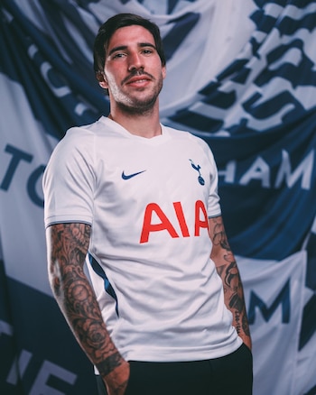 Hombre joven con barba, camiseta blanca de fútbol con el escudo del Tottenham y logo de Nike y AIA, brazos tatuados, fondo con bandera del equipo