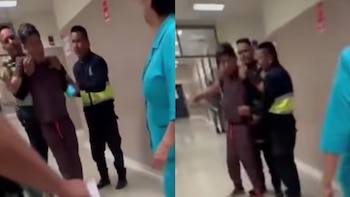 Tres personas en un pasillo de hospital. Un hombre es sujetado por dos personas, una de ellas con chaleco reflectante. Otra persona se ve parcialmente