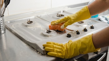 Manos con guantes amarillos limpian una estufa de cuatro quemadores con una esponja naranja. Utensilios de cocina se ven en un recipiente metálico.