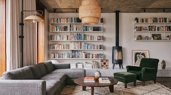 Salón moderno con sofá gris, sillón verde con reposapiés, mesa de centro de madera, estanterías blancas con libros, chimenea negra y lámpara colgante de mimbre.