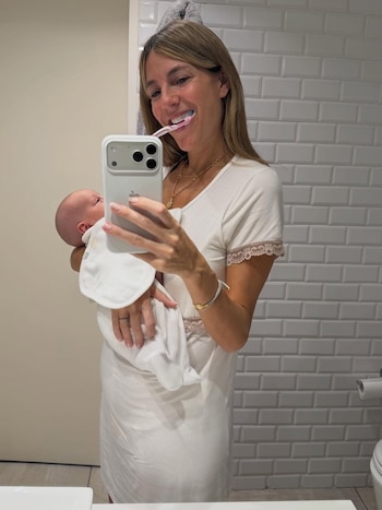 Una mujer con cabello castaño, vestida de blanco, se toma una selfie en el espejo del baño, cepillándose los dientes mientras sostiene a un bebé envuelto en blanco