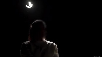 Una persona sentada, con la espalda hacia el espectador, está silueteada por una luz brillante desde arriba en un fondo oscuro