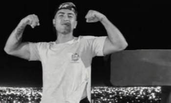 Fotografía en blanco y negro de un hombre joven que flexiona los brazos, viste gorra y camiseta, con las luces de la ciudad de fondo