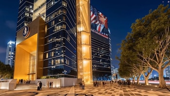 Edificio moderno iluminado de noche con una gran pantalla digital mostrando a Donald Trump y la bandera de EE. UU. en su fachada. Personas transitan en la plaza adyacente