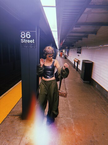 Una mujer con lentes y ropa verde posa junto a un pilar con el cartel 86 Street en una estación de metro