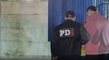 Vista trasera de un oficial de PDI con chaqueta negra y un hombre con sudadera rosa, caminando frente a una pared clara y una estructura azul