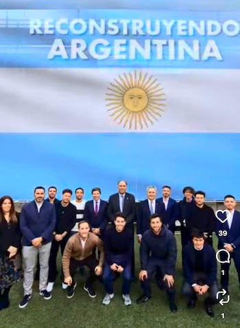 Un grupo de hombres, algunos de pie y otros agachados, posan frente a un banner con la bandera argentina y la frase "Reconstruyendo Argentina"