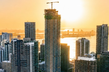 Vista aérea de un paisaje urbano con múltiples rascacielos. Un edificio central en construcción con grúa, bahía y el sol reflejado en el horizonte