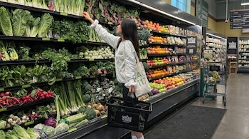 Una mujer en un supermercado Whole Foods Market selecciona productos frescos de estantes llenos de vegetales y frutas, sosteniendo una cesta de compras.