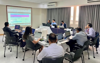 Nueve personas sentadas en mesas con laptops abiertas en una sala de conferencias, con una hoja de cálculo de Excel proyectada en una pantalla