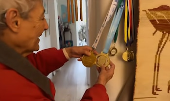 Una mujer mayor de perfil sonríe mirando sus manos que sostienen dos medallas doradas, con más medallas colgando en la pared de una habitación junto a un tapiz