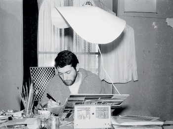 Fotografía en blanco y negro de Julio Le Parc, un hombre con barba sentado en un escritorio lleno de herramientas y materiales, bajo una lámpara