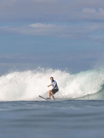 Hombre surfeando una gran ola en una tabla Wavestorm negra. Viste una camiseta azul y blanca y pantalones cortos negros. Cielo azul con nubes