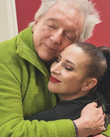 Un hombre y una mujer se abrazan. El hombre tiene pelo gris y chaqueta verde. La mujer tiene pelo oscuro y labial rojo. Ambos tienen los ojos cerrados