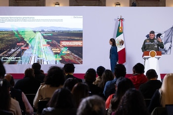Un militar habla en un podio junto a Claudia Sheinbaum ante una pantalla con un mapa de rutas ferroviarias y un público de espaldas