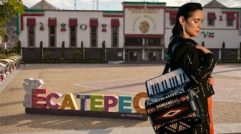 Una mujer sostiene un acordeón negro frente a las letras decorativas de Ecatepec y el palacio municipal.