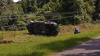 Una SUV oscura está volcada sobre su techo en una zanja cubierta de hierba al lado de una carretera, con un oficial de policía agachado cerca