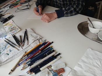 Vista cenital de Enrique Breccia dibujando con un lápiz azul sobre papel blanco. La mesa está llena de lápices de colores, pinceles, borradores y un trapo con manchas de pintura