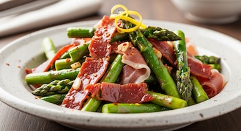 Un plato hondo contiene espárragos verdes, láminas de jamón serrano, ralladura de limón, hojuelas de chile seco y trozos de ajo.