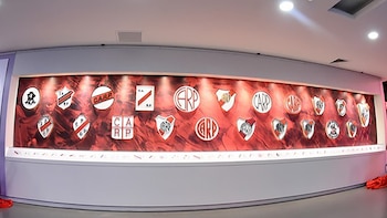 Vista panorámica de una sala del Museo River con una exhibición cronológica de los escudos de River Plate en una pared roja iluminada, con personas observando