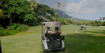 Dos carros de golf en un campo de golf verde con palmeras, vegetación tropical densa, un cuerpo de agua pequeño y una montaña al fondo bajo un cielo nublado