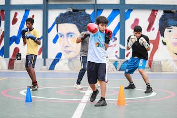Cuatro jóvenes entrenando boxeo al aire libre en una cancha. El niño central lleva guantes rojos y simula un golpe. Un mural con figuras humanas adorna la pared trasera