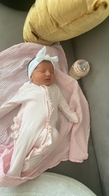 Una bebé recién nacida con un mono rosa de punto y un lazo blanco en la cabeza duerme sobre una manta rosa, junto a un biberón y un cojín dorado