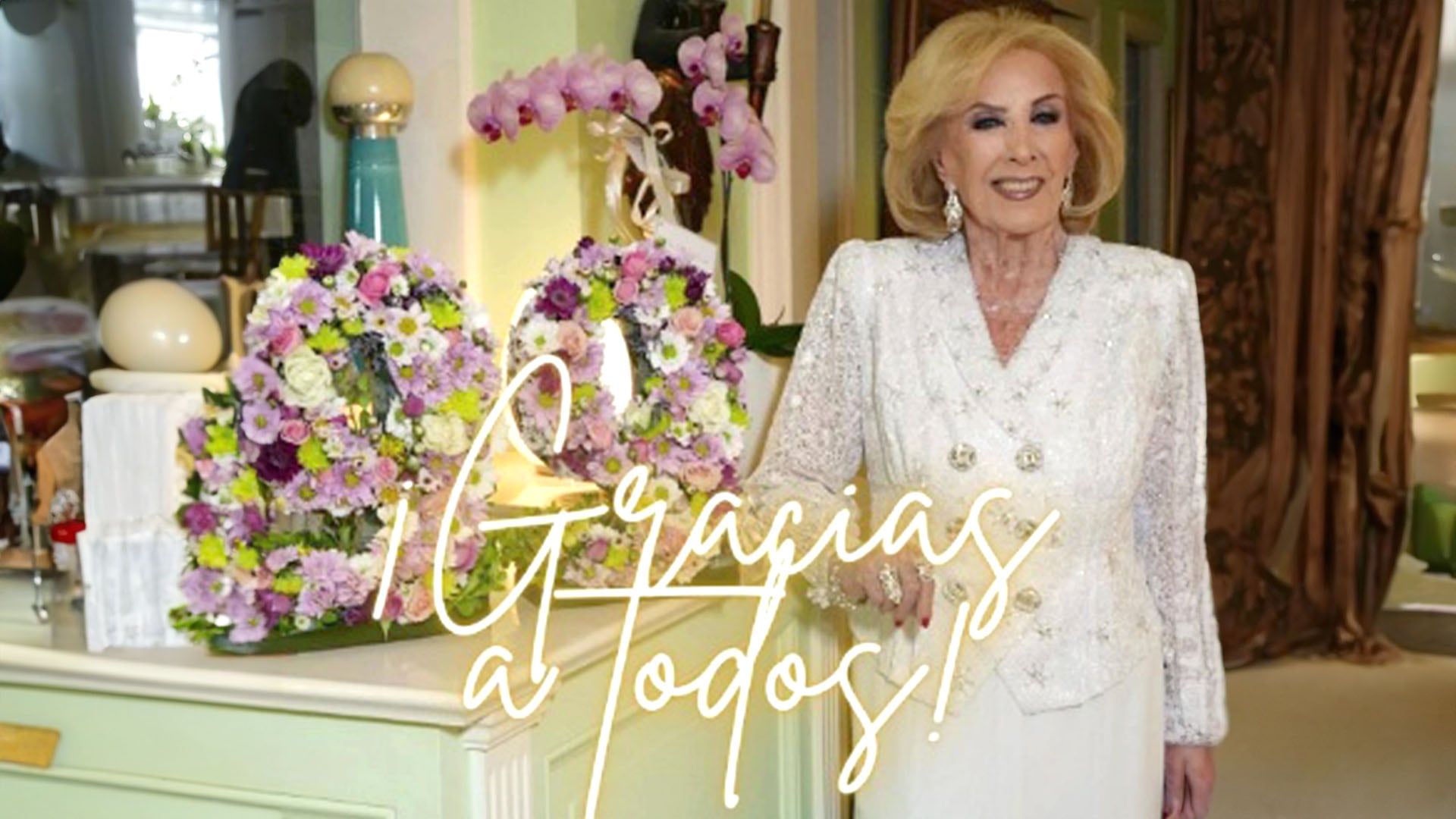 La icónica presentadora Mirtha Legrand posa sonriente junto a dos exuberantes arreglos florales, expresando su gratitud en su día de cumpleaños. (Captura de video)