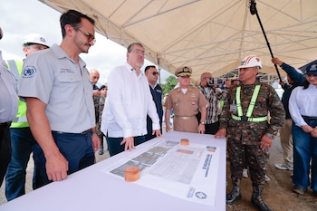 Hombres con camisas y uniformes militares observan planos sobre una mesa, bajo una carpa blanca al aire libre