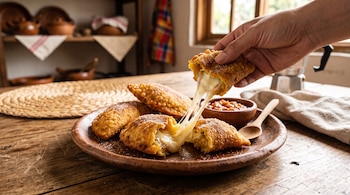 Empanadas fritas en un plato de barro sobre una mesa de madera y una mano que levanta media pieza con queso derretido estirado.