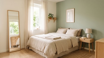Dormitorio con cama de lino beige, mesa de noche con lámpara, espejo de cuerpo entero, ventana y pared verde salvia.