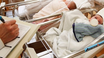 Primer plano de una mano escribiendo en un portapapeles junto a dos bebés recién nacidos en cunas de hospital; uno de ellos toma un biberón