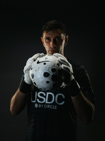 Emiliano Martínez sostiene un balón de fútbol con ambos guantes blancos sobre un fondo oscuro