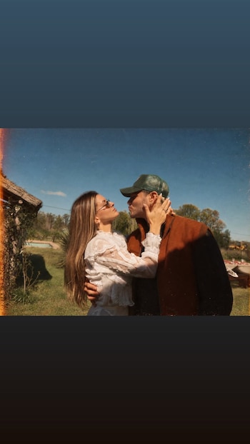 Una mujer con camisa blanca de encaje y gafas de sol abraza a un hombre con gorra verde y chaleco marrón en un campo soleado