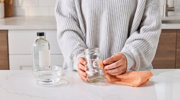 Una persona usa un paño naranja para limpiar un frasco de vidrio con residuos de papel, junto a una botella y un cuenco con líquido.