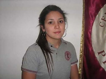 Una mujer joven con cabello oscuro y camisa polo gris posa de frente junto a una bandera bordada