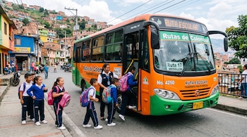 Niños con uniforme escolar suben a un autobús naranja y verde acompañados por una mujer en una calle urbana junto a casas en la colina.