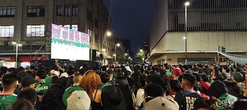 Multitud de personas vista desde atrás observa una pantalla gigante en la calle, que muestra un partido de fútbol. Edificios y farolas a los lados por la noche