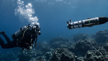 Buzo con traje negro, equipo de buceo y burbujas ascendiendo. A su lado, vehículo submarino autónomo cilíndrico con franjas y luces verdes. Fondo marino rocoso