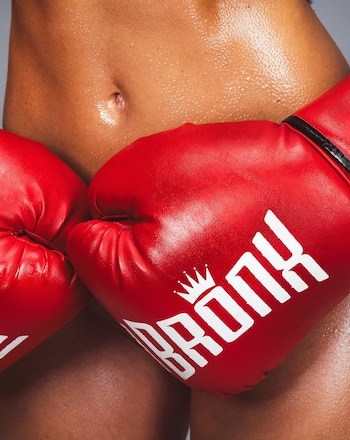 Plano detalle del abdomen transpirado de una persona que sostiene dos guantes de boxeo rojos con la marca Bronx