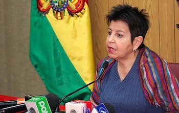 Teresa Morales habla frente a varios micrófonos de prensa junto a una bandera boliviana